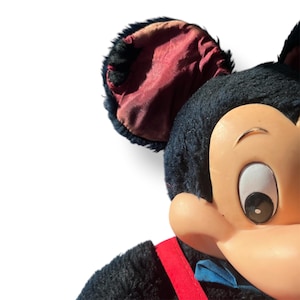 May include: Close-up of a vintage Mickey Mouse plush toy. The plush features a black body, a tan face, and red suspenders. The ears are black with a burgundy interior. A blue bow tie adds a pop of color.