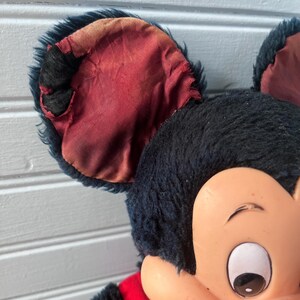 May include: Close-up of a vintage Mickey Mouse plush toy. The toy features black fur, large ears with red lining, and a plastic face with a cheerful expression. The plush has a red outfit.