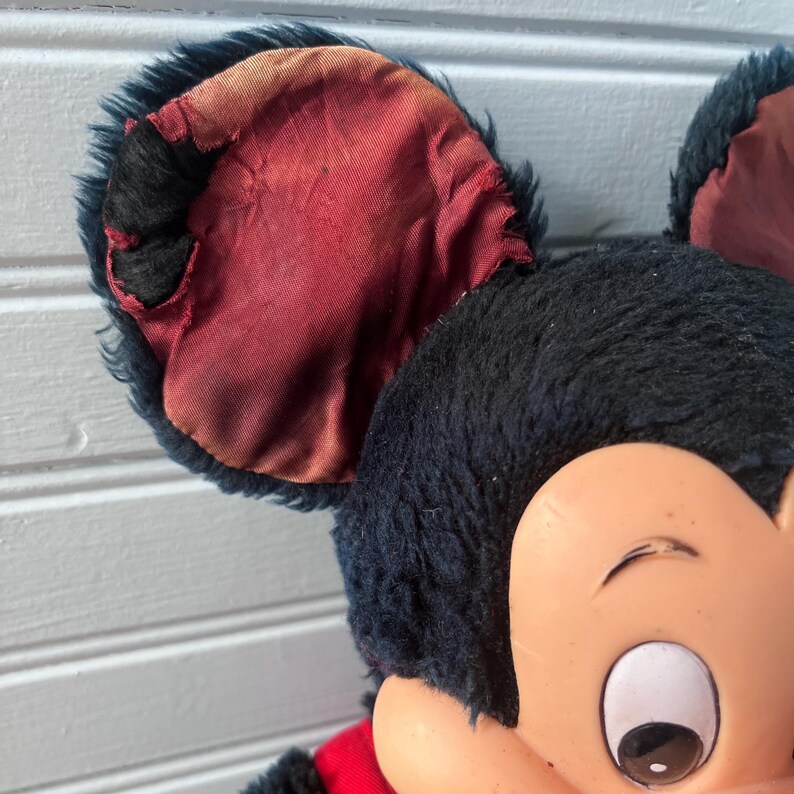 May include: Close-up of a vintage Mickey Mouse plush toy. The toy features black fur, large ears with red lining, and a plastic face with a cheerful expression. The plush has a red outfit.