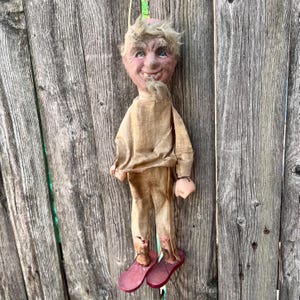 May include: A hanging doll with a textured, tan-colored fabric outfit, featuring a sculpted face with a wide, toothy grin, light-colored hair, and a small goatee. The doll wears flat, red-painted wooden shoes and has visible dark stains on the pant legs.