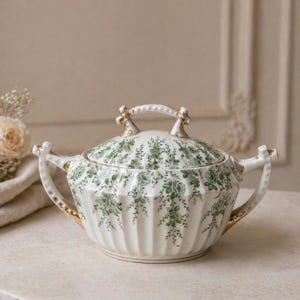 Może przedstawiać: Biała porcelanowa salaterka z pokrywką, ozdobiona zielonym wzorem kwiatowym. Naczynie ma żebrowany wzór i złote akcenty na uchwytach i pokrywce. Uchwyty są zakrzywione, a pokrywka ma uchwyt pętelkowy.