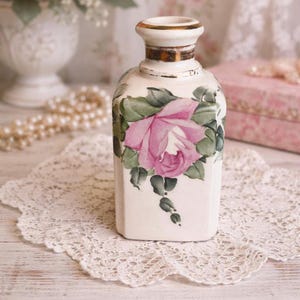 Puede incluir: Un jarrón rectangular de cerámica vintage con una rosa rosa pintada a mano y hojas verdes. El jarrón tiene un borde dorado y está sobre un tapete de encaje blanco. El fondo incluye un jarrón blanco borroso y perlas.