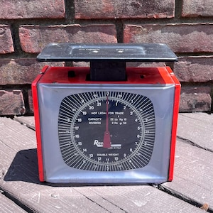 May include: A red and black mechanical kitchen scale with a square face featuring a black dial, white markings, and a red needle. The dial displays text including Capacity 22 Lb, Division 2 Oz, and Not Legal For Trade. It has a black top platform.