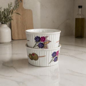 May include: Two white ceramic ramekins stacked on top of each other. Each ramekin has a ribbed design and is decorated with illustrations of olives and berries in shades of purple, pink, and green.