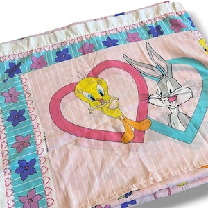 Könnte beinhalten: Rosa-weißes Looney Tunes Bettlaken mit Tweety und Bugs Bunny in herzförmigen Rahmen. Das Laken hat ein gestreiftes Muster mit floralen und herzförmigen Akzenten in Blau, Rosa und Lila.