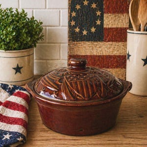 Könnte beinhalten: Eine braune Keramik-Auflaufform mit Deckel, der ein geprägtes amerikanisches Flaggenmuster aufweist. Die Form hat Griffe und steht auf einer Holzoberfläche. Ein patriotisches Thema ist mit einer Flagge und sternverzierten Gegenständen im Hintergrund vorhanden.