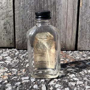May include: A small, clear glass bottle with a black cap. The bottle has a paper label with faded text. The bottle is sitting on a rough, gray surface in front of a weathered wooden background. The bottle contains a liquid.
