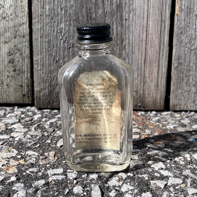 May include: A small, clear glass bottle with a black cap. The bottle has a paper label with faded text. The bottle is sitting on a rough, gray surface in front of a weathered wooden background. The bottle contains a liquid.