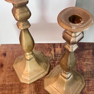 May include: Two antique brass candlesticks. Each candlestick has a hexagonal base, a decorative stem, and a circular top with a hole for a candle. The candlesticks are a warm, golden color and appear to be made of metal.
