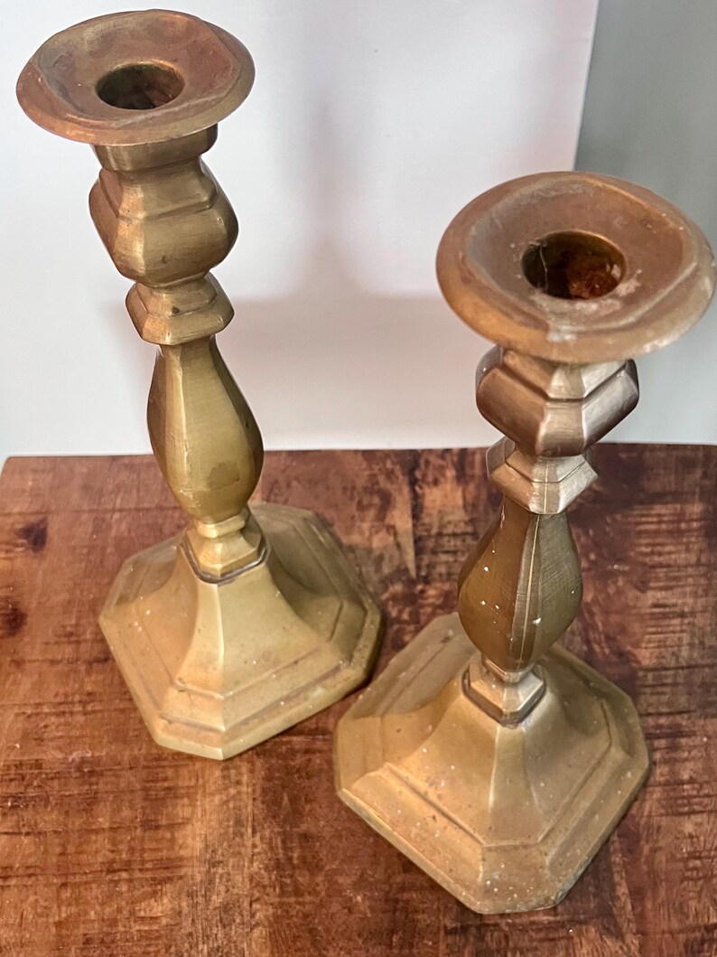 May include: Two antique brass candlesticks. Each candlestick has a hexagonal base, a decorative stem, and a circular top with a hole for a candle. The candlesticks are a warm, golden color and appear to be made of metal.