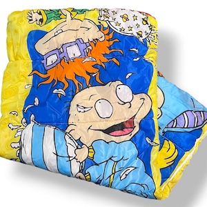May include: A colourful, quilted sleeping bag featuring cartoon characters in a vibrant design. The bag has a yellow border and a blue and yellow interior with cartoon graphics. The design includes characters with orange hair and purple glasses.