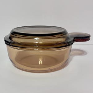May include: A vintage amber-colored glass casserole dish with a matching lid. The dish has a single handle and a rounded shape. The glass is transparent, allowing the contents to be seen.