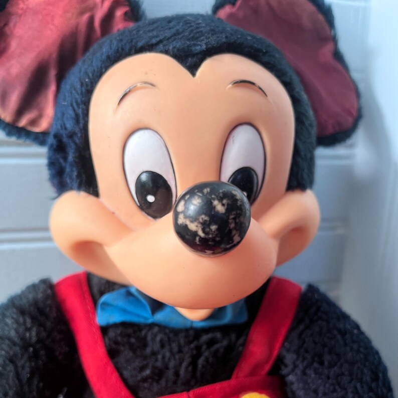May include: Close-up of a Mickey Mouse plush toy. The toy has a black, plush head with large, white-rimmed eyes and a black nose. It wears a red bow tie and red suspenders over a black body. The ears are a dark red.