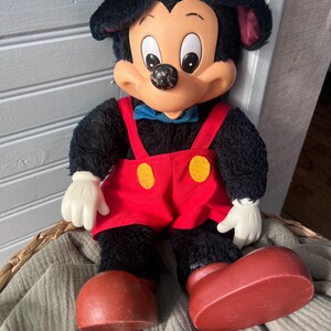 May include: A vintage Mickey Mouse plush doll. The doll has a black body, large red shoes, and red overalls with yellow buttons. It has a white face, hands, and large ears. A blue bow tie completes the look.
