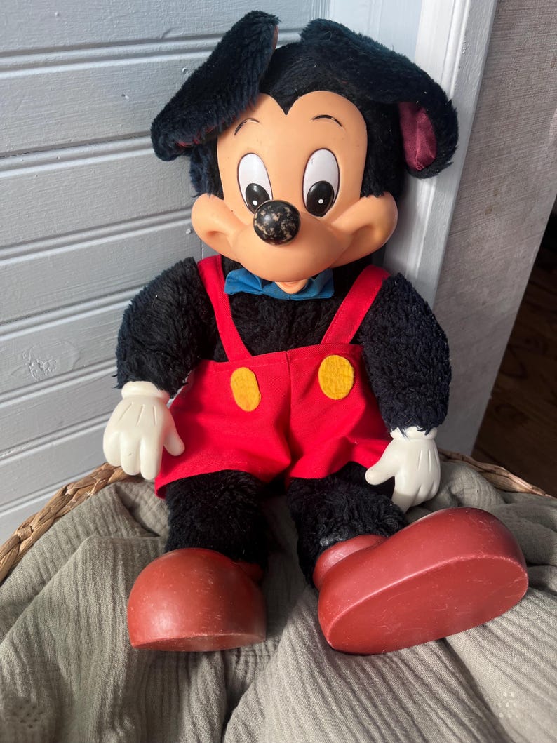 May include: A vintage Mickey Mouse plush doll. The doll has a black body, large red shoes, and red overalls with yellow buttons. It has a white face, hands, and large ears. A blue bow tie completes the look.