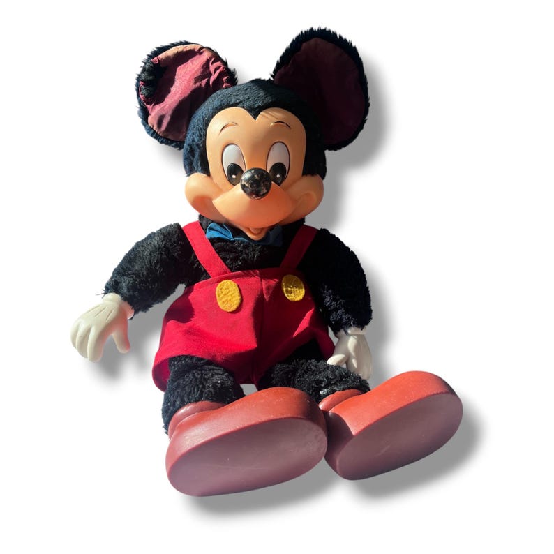 May include: A plush Mickey Mouse doll with black fur, large ears, and a smiling face. He wears red overalls with yellow buttons, red shoes, white gloves, and a blue bow tie. The doll is sitting down.