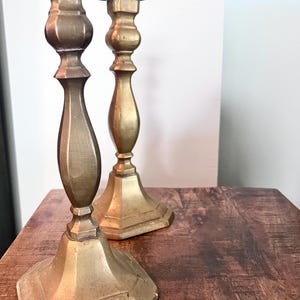 May include: Two antique brass candlesticks with a warm, golden hue. The candlesticks have a classic design with a wide, octagonal base, a slender stem, and a flared top. They are placed on a wooden surface, showcasing their vintage appeal.