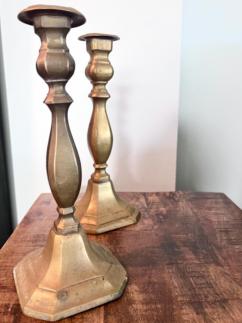 May include: Two antique brass candlesticks with a warm, golden hue. The candlesticks have a classic design with a wide, octagonal base, a slender stem, and a flared top. They are placed on a wooden surface, showcasing their vintage appeal.