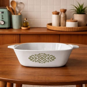 May include: White rectangular casserole dish with a green and gold decorative pattern. The dish has handles on each side and is sitting on a wooden table. Kitchenware.
