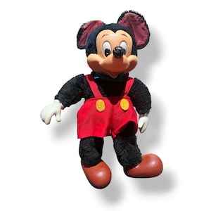 May include: A vintage Mickey Mouse plush doll. The doll features a black body, red shorts with yellow buttons, white gloves, and brown shoes. Mickey's face is tan with a black nose and large, round eyes. The doll has large, black ears and a blue bow tie.
