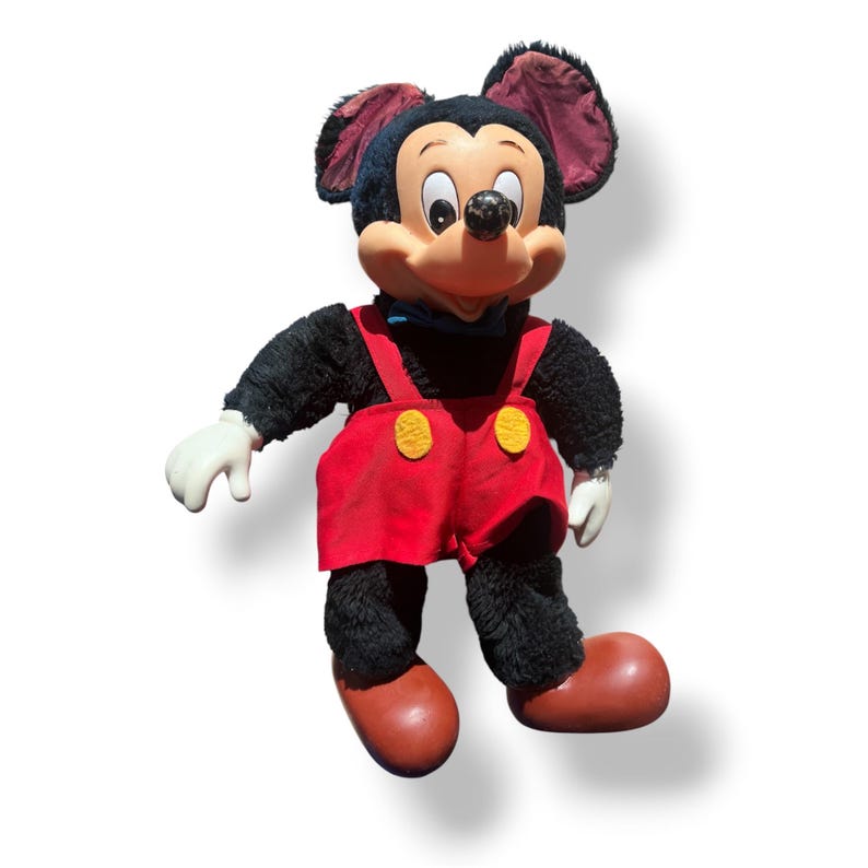 May include: A vintage Mickey Mouse plush doll. The doll features a black body, red shorts with yellow buttons, white gloves, and brown shoes. Mickey's face is tan with a black nose and large, round eyes. The doll has large, black ears and a blue bow tie.