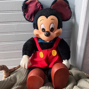 May include: A vintage Mickey Mouse plush doll. The doll has black fur, large ears with burgundy lining, and a smiling face. It wears red overalls with yellow buttons, white gloves, and large brown shoes. A blue bow tie completes the look.