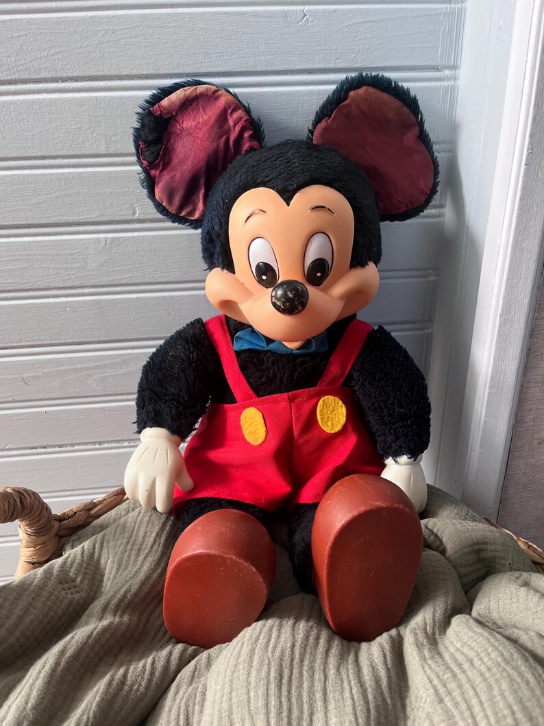 May include: A vintage Mickey Mouse plush doll. The doll has black fur, large ears with burgundy lining, and a smiling face. It wears red overalls with yellow buttons, white gloves, and large brown shoes. A blue bow tie completes the look.