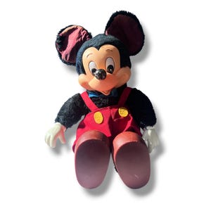 May include: A vintage Mickey Mouse plush doll. The doll has a black body, large ears, and a smiling face. It wears red overalls with yellow buttons, white gloves, and brown shoes. A blue bow tie completes the classic look.