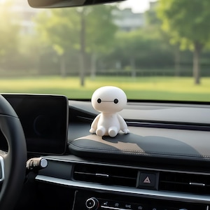 May include: A white, cartoon-style figurine with a rounded head and body, black eyes, and a horizontal black line for a mouth. The figurine is sitting on a car dashboard, with a blurred background of trees and sunlight.