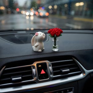 May include: A white ceramic bunny figurine with a pink flower on its head sits next to a small glass vase holding red flowers. The items are on a black dashboard, with a blurred street scene in the background.