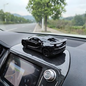May include: A black toy car with a spoiler sits on a dashboard. The car has a sleek design with visible details like the wheels and windows. The dashboard has a carbon fiber pattern.
