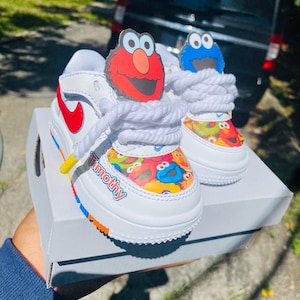 May include: White toddler sneakers with red and blue accents, featuring Sesame Street character designs. The shoes have white laces and the name "Timothy" printed on the side. The sneakers are on a white box.