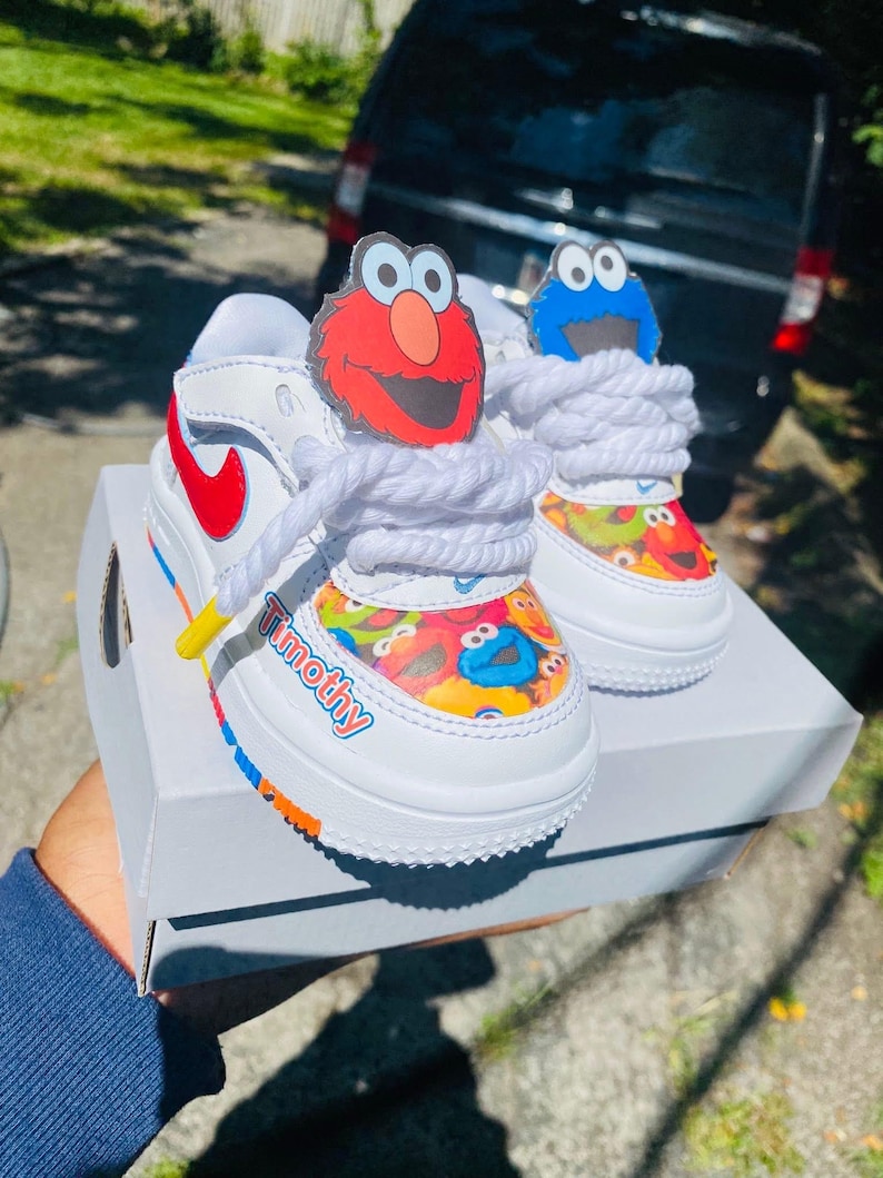 May include: White toddler sneakers with red and blue accents, featuring Sesame Street character designs. The shoes have white laces and the name "Timothy" printed on the side. The sneakers are on a white box.