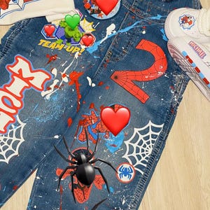 May include: A denim overall set with a matching t-shirt and sneakers. The overalls and t-shirt feature a Spider-Man theme with the number "2", spiderwebs, and the text "HAEL" and "TEAM UP!". The sneakers have the name "MICHAEL" on them.