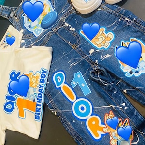 May include: A denim outfit set for a child, featuring a white t-shirt with "Birthday Boy" text and a number 1 graphic, denim overalls, and jeans with character graphics and paint splatter details. White and blue sneakers complete the ensemble.