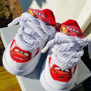 May include: White sneakers featuring a red Lightning McQueen graphic on the toe and tongue. The shoes have thick white laces and a red accent on the heel. The shoes are sitting on a gray surface.