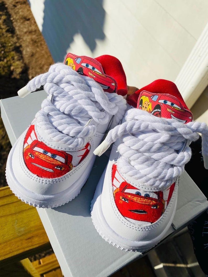May include: White sneakers featuring a red Lightning McQueen graphic on the toe and tongue. The shoes have thick white laces and a red accent on the heel. The shoes are sitting on a gray surface.