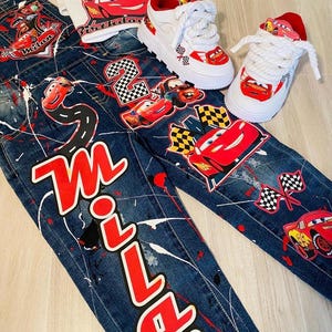 May include: A set of children's clothing featuring a Cars theme. Includes denim overalls and a t-shirt with the name "Milan" and Lightning McQueen graphics. White sneakers with red accents and Cars details complete the ensemble.