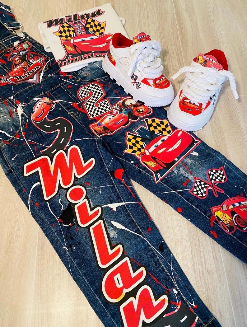 May include: A set of children's clothing featuring a Cars theme. Includes denim overalls and a t-shirt with the name "Milan" and Lightning McQueen graphics. White sneakers with red accents and Cars details complete the ensemble.