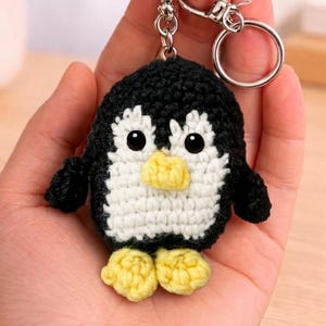 Puede incluir: Un llavero de pingüino tejido a mano. El pingüino tiene un cuerpo negro, vientre blanco, pico y pies amarillos y ojos negros. El llavero incluye un cierre y un anillo plateados.