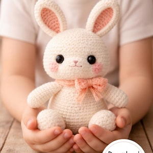 May include: A crocheted bunny doll with a cream-colored body, pink inner ears, and rosy cheeks. It has black eyes and a pink bow tie. The doll is held in someone's hands. A button in the bottom right corner says "Download PDF Pattern".