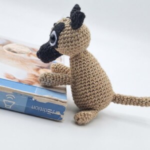 May include: A small, handmade crochet dog figurine crafted from tan yarn with a black muzzle, black ears, and black patches around the eyes. The toy features large, round plastic eyes and a long, thin tail, standing upright on a plain white surface.