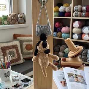 May include: A display featuring a crocheted dog and a small gray crocheted purse hanging from a wooden board. The dog is tan with black accents. The board has a sign that reads "Amigurumi-Träume". Shelves of yarn are in the background.