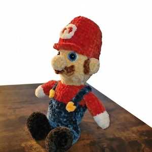 Könnte beinhalten: Handgemachte Häkelpuppe der Videospiel-Figur Mario. Die Puppe hat einen roten Hut mit weißem Emblem, ein rotes Hemd, blaue Latzhosen und schwarze Schuhe. Die Puppe hat ein beiges Gesicht, einen braunen Schnurrbart und blaue Augen.