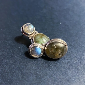 Puede incluir: Un anillo plateado con cuatro piedras de labradorita ovaladas y redondas. Las piedras muestran tonos azules, verdes y grises. El anillo tiene un diseño inusual y orgánico.