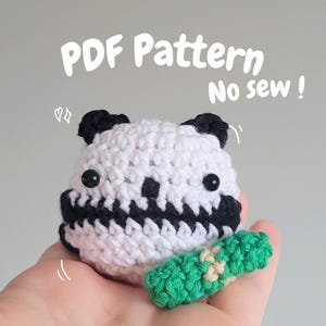 Pode incluir: Um urso panda de crochê branco e preto com olhos de botão pretos e um pequeno acessório verde e bege. A imagem inclui o texto "PDF Pattern No sew!" Feito à mão.