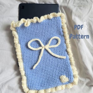 Puede incluir: Una funda de tableta tejida a ganchillo en color azul claro con un borde de volantes color crema y un lazo central color crema. Un pequeño corazón color crema está cosido en la esquina inferior derecha. El texto PDF Pattern es visible.