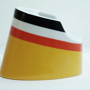 May include: A ceramic ashtray with a distinctive design. The ashtray features a yellow base, a red stripe, a white stripe, and a black top. The design is reminiscent of a flag. The ashtray has a curved shape and a small indentation for resting cigarettes.