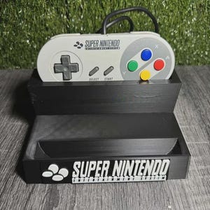 Puede incluir: Un soporte negro para mando de Super Nintendo con un mando a juego. El soporte tiene las palabras "SUPER NINTENDO ENTERTAINMENT SYSTEM" en blanco. El mando es gris con botones de colores y una cruceta.