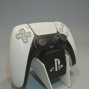Puede incluir: Un mando de videojuegos blanco y negro con un soporte. El mando tiene un diseño simétrico con botones y joysticks. El soporte es blanco con un panel negro con un logotipo.